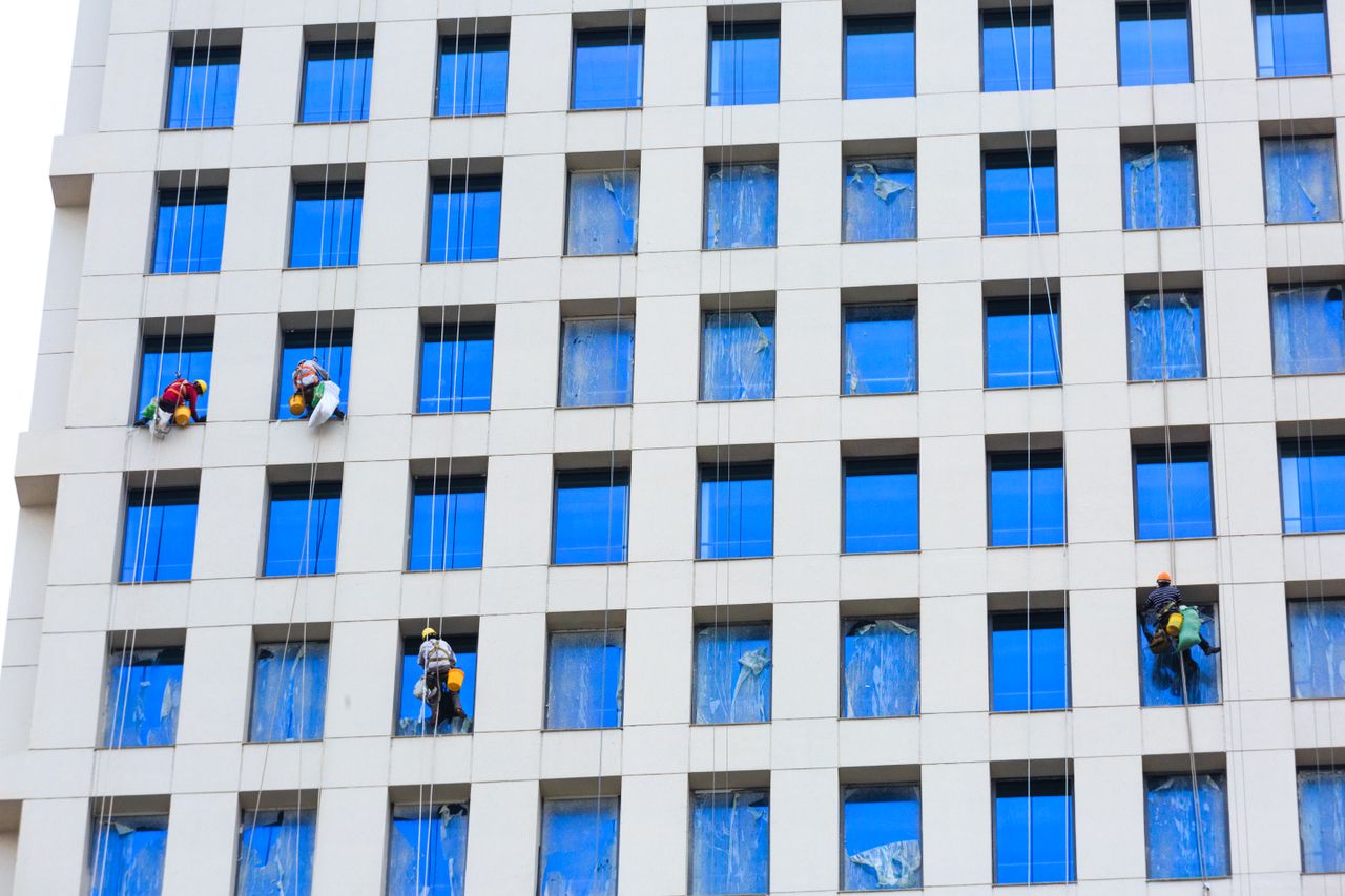 Des professionnels nettoient les vitres d’un gratte-ciel à l’aide de cordes, dans un environnement urbain.